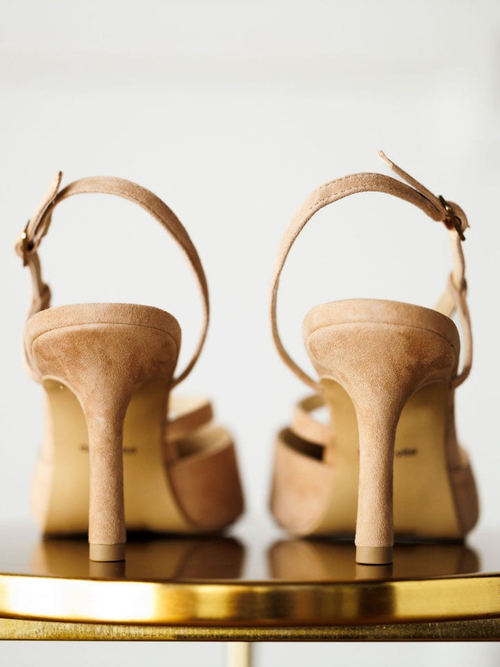 A close-up of the back of The Lafayette beige high-heeled sandals by Savannah Sevens western life{&}style, featuring ankle straps with adjustable buckles, displayed on a shiny gold surface against a light background.
