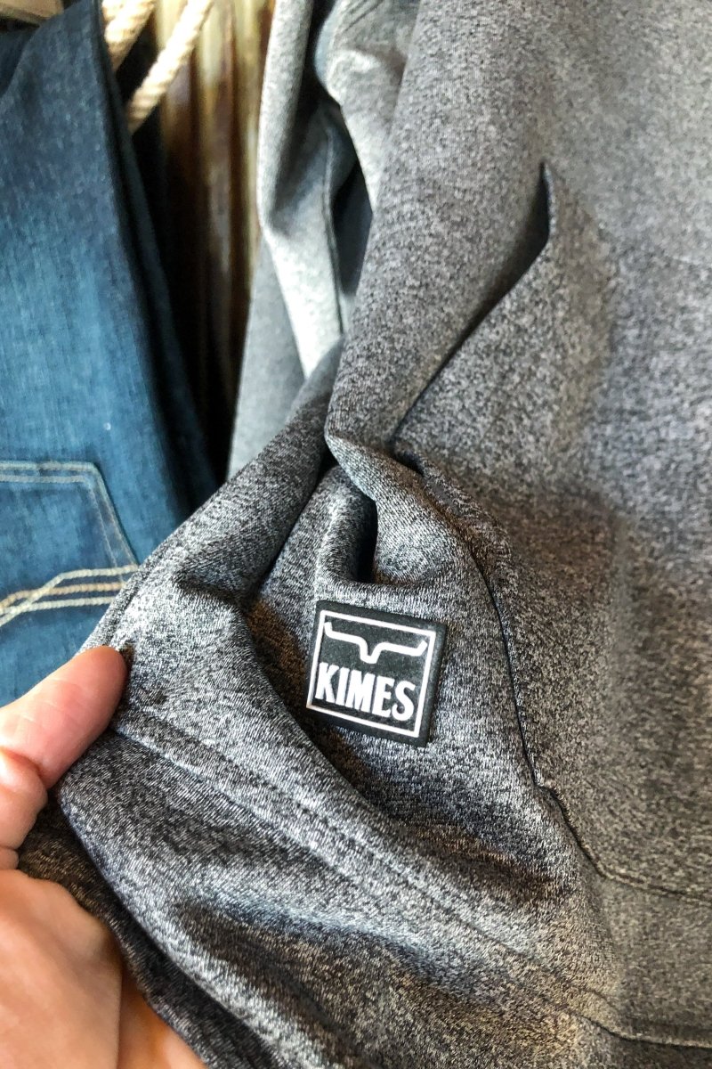 A hand holds The Lanesboro {Men's} gray tech fabric hoodie from Savannah Sevens western life{&}style, featuring a stitched black and white logo tag. Blue denim with visible stitching appears in the background.