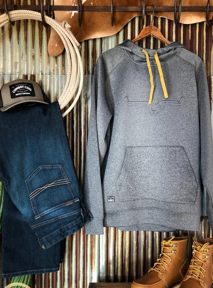 The Lanesboro hoodie by Savannah Sevens western life{&}style, featuring gray fabric and yellow drawstrings, is styled with dark blue jeans, a black trucker cap, and tan work boots against a corrugated metal wall with wooden hooks above.