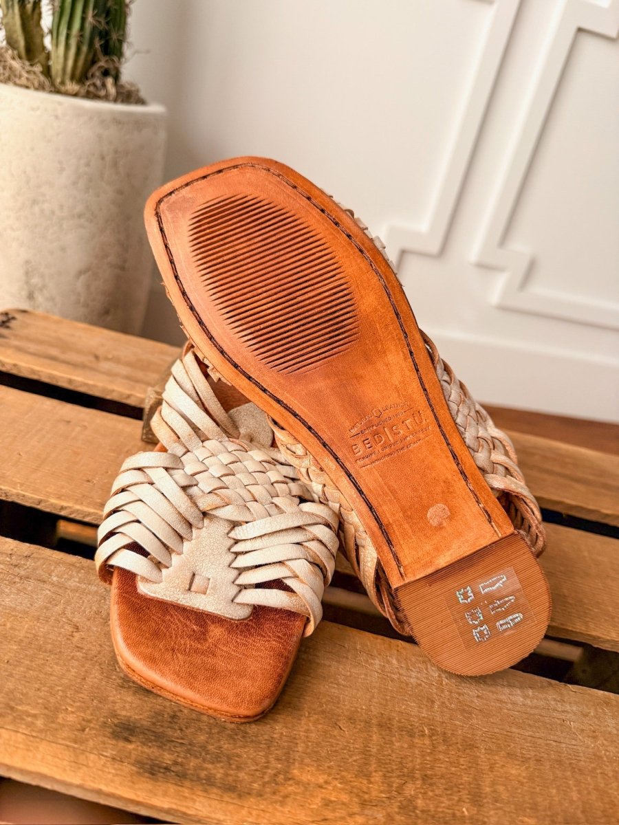 The Layton by Savannah Sevens western life{&}style: hand woven leather open toe flat sandals, displayed on wood—one highlights the detailed sole, while the other showcases the intricate braided design on top. Sandals made by BedStu. 