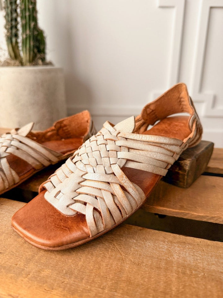 A close-up of The Layton huarache sandal by Savannah Sevens western life{&}style, featuring hand-woven leather straps on a wooden surface, with another sandal and a potted cactus softly blurred in the background.