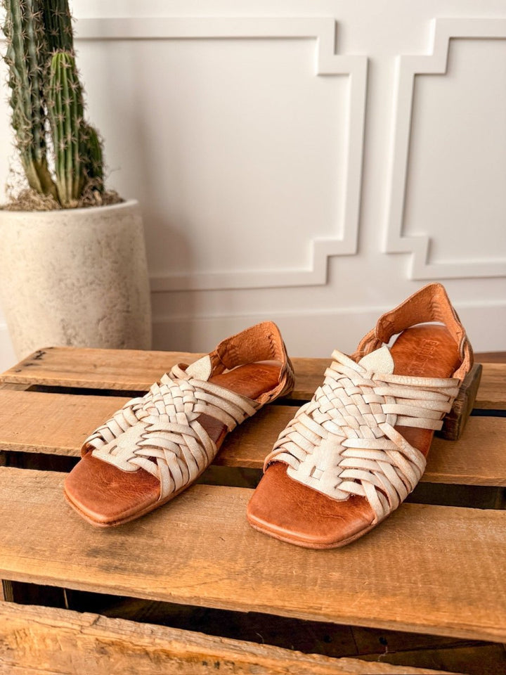 The Layton handwoven leather sandals in tan and white by Savannah Sevens western life{&}style rest on a wooden pallet, with a tall cactus in a textured planter against a decorative white wall in the background. Sandals by Bed Stu.