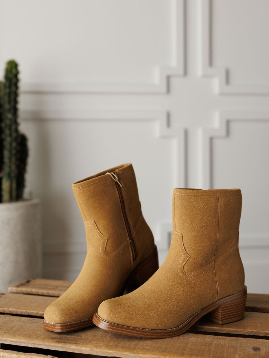 The Linden by Savannah Sevens western life{&}style features tan square toe bootie ankle boots with low wooden heels, displayed on a wooden surface with a potted cactus and a white wall with decorative molding in the background.