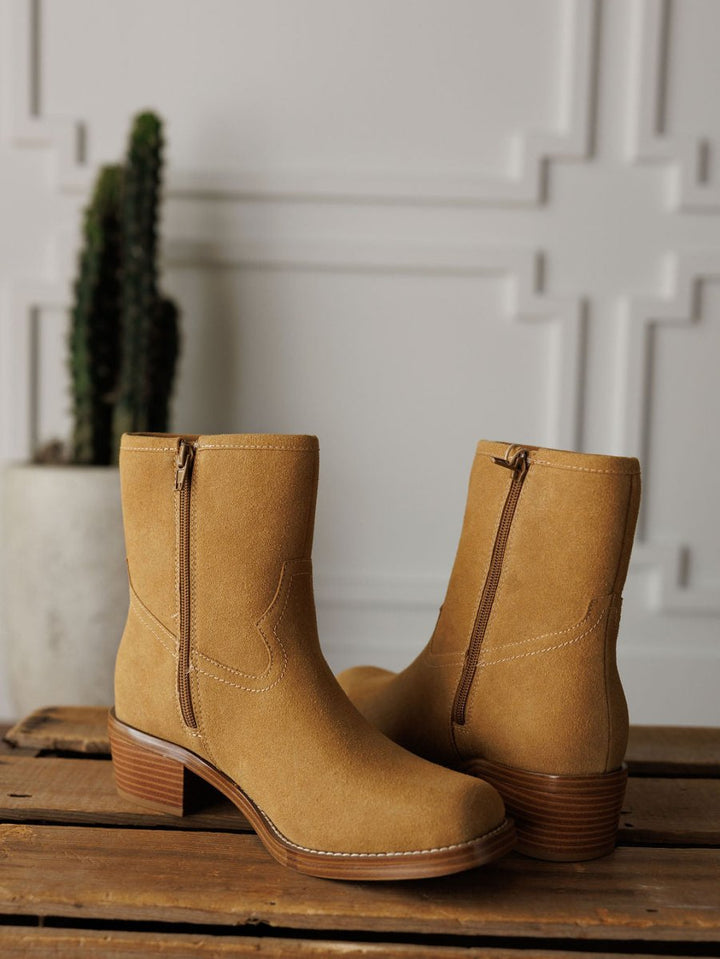 The Linden bootie by Savannah Sevens western life{&}style features tan leather, side zippers, and stacked wooden heels. Displayed on rustic wood with a potted cactus and white decorative wall accentuating its western style.