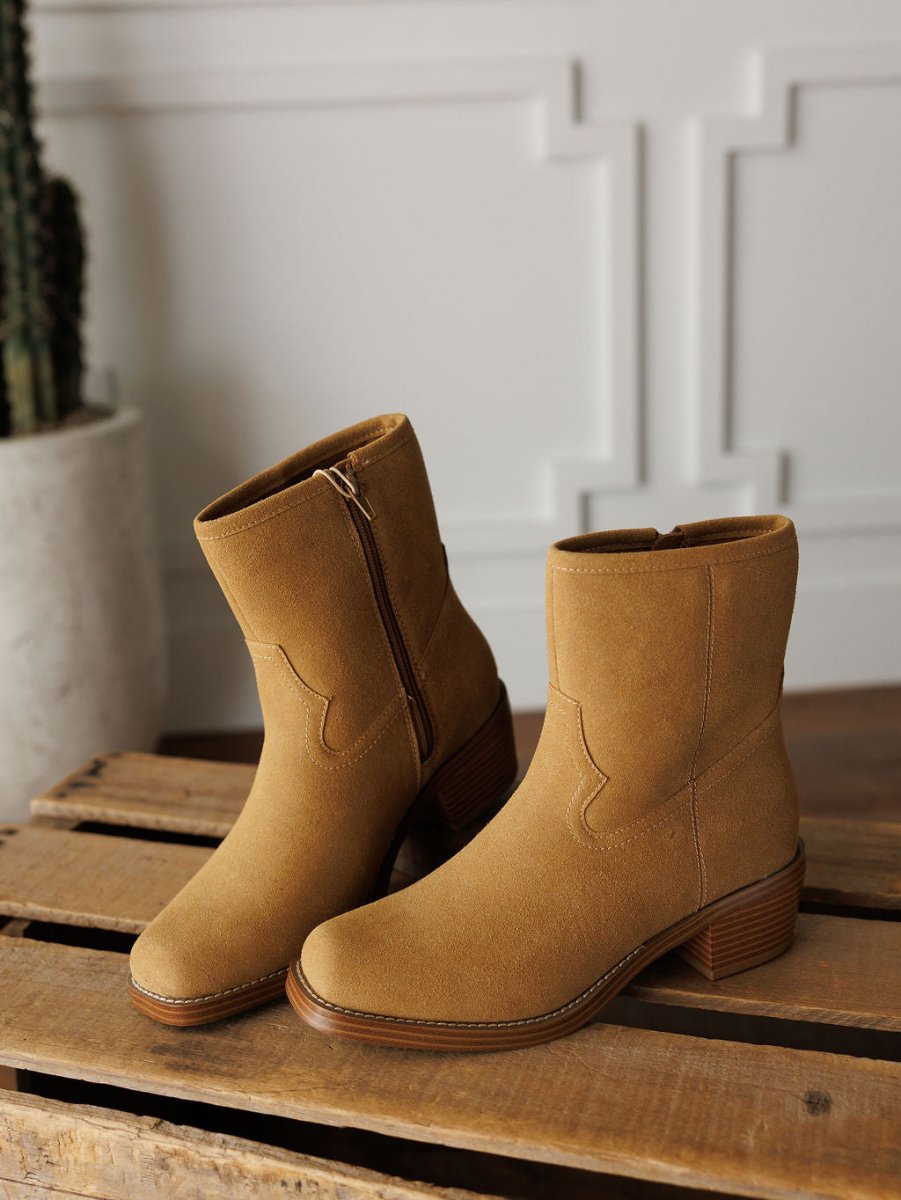 The Linden booties by Savannah Sevens western life{&}style, in tan suede with low stacked heels and side zippers, are shown on a wooden crate with a potted cactus and white paneled walls in the background.