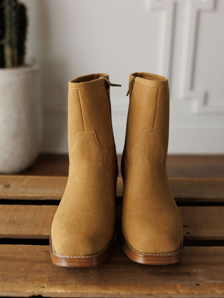 The Linden booties by Savannah Sevens western life{&}style, crafted from tan leather with side zippers, rest on a wooden surface. A blurred potted cactus and white wall paneling can be seen in the background.