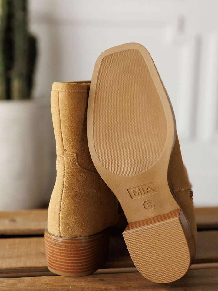 A close-up of The Linden tan suede western booties by Savannah Sevens western life{&}style on wood, one upright and one displaying its sole with size 8. A blurred potted plant appears in the background.