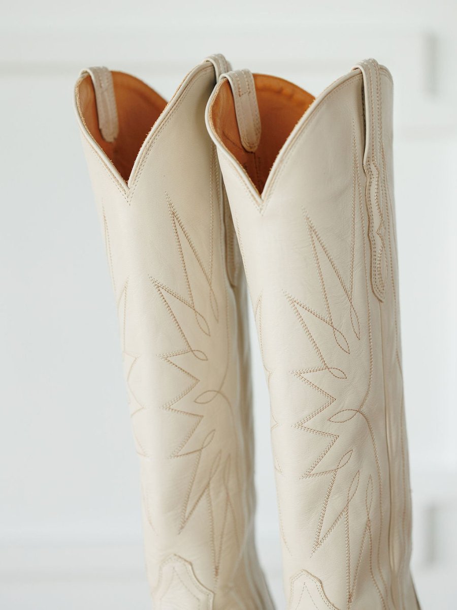 A close-up of The Loretta Boot {Cream} by Savannah Sevens western life{&}style, tall snip toe boots in light beige premium cowhide leather with decorative stitching and pull tabs, set against a white background. Boots handmade by Tecovas.