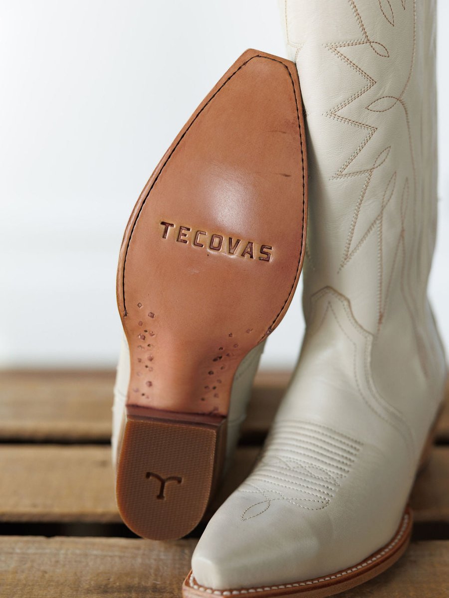 A close-up of the sole of a cream The Loretta Boot by Savannah Sevens western life{&}style, crafted from premium leather, resting on wood with its matching pair in the background. Boots handmade by Tecovas.