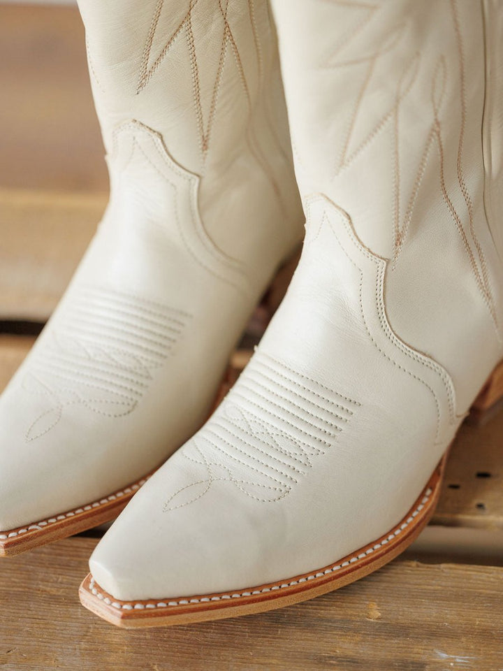 Close-up of The Loretta Boot {Cream} by Savannah Sevens western life{&}style—stylish white cowboy boots with decorative stitching, pointed toes, tan soles, and premium cowhide leather, set on a wooden surface. Boots handmade by Tecovas.