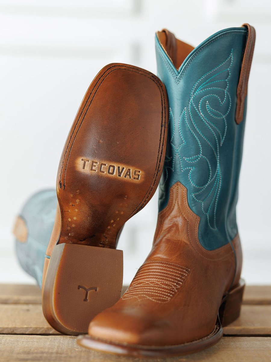 A pair of The Maggie Boot {Teal} by Savannah Sevens western life{&}style with tan leather feet, turquoise shafts, classic Western square toe, and decorative stitching—one standing upright, the other tilted to show the embossed sole on wood. Boots handmade by Tecovas.