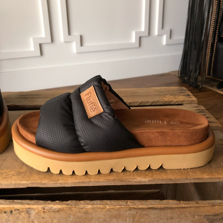 A close-up of The Marfa {Black} puffer sandal by Savannah Sevens western life{&}style, featuring a thick tan sole and cushioned insole, displayed on rustic wood with a white paneled wall in the background. Sandals made by Flufie. 