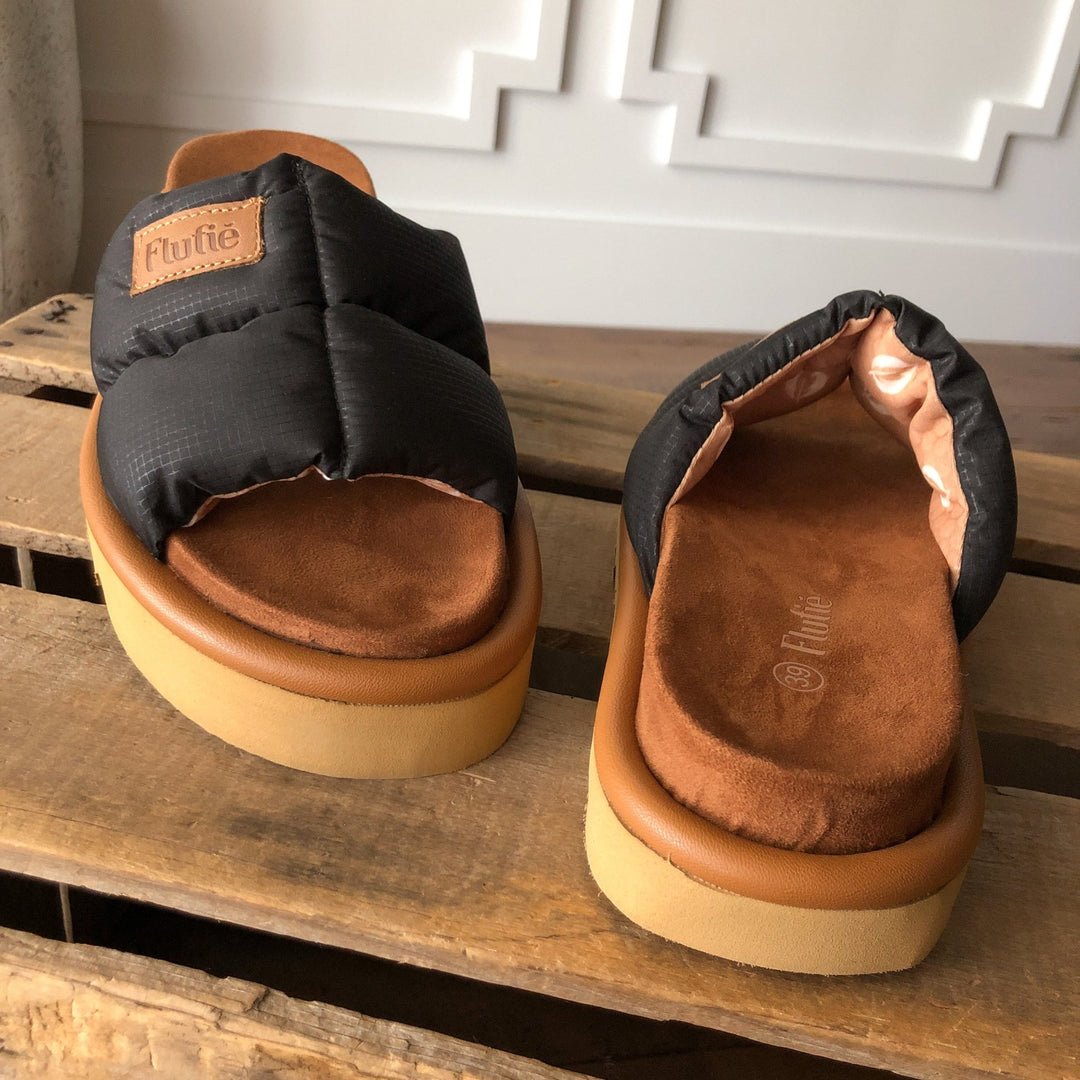 The Marfa {Black} puffer sandals by Savannah Sevens western life{&}style, featuring cushioned insoles and thick tan shock-absorbing outsoles, are displayed on a rustic wooden surface indoors. Sandals by Flufie. 