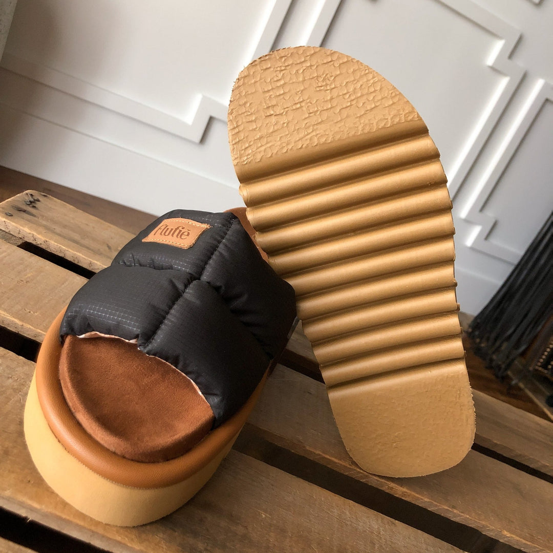 A close-up of The Marfa {Black} puffer sandals by Savannah Sevens western life{&}style shows one with a shock-absorbing outsole, the other with a cushioned insole and brand label, resting on an indoor wooden surface.