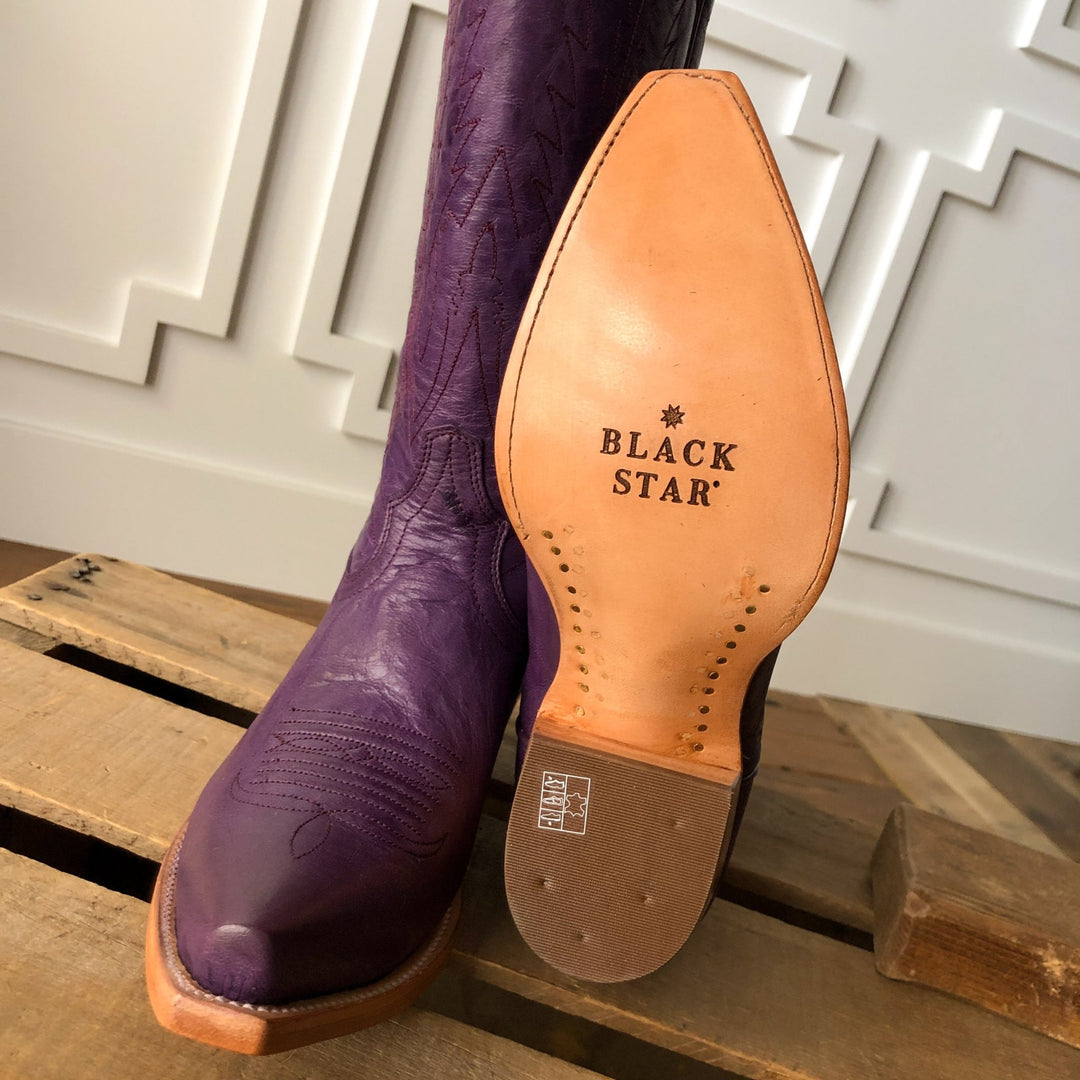 A close-up of The Maricopa by Savannah Sevens western life{&}style shows a plum leather snip toe boot and another with its tan sole stamped, both displayed on a wooden surface. Made by Black Star. 