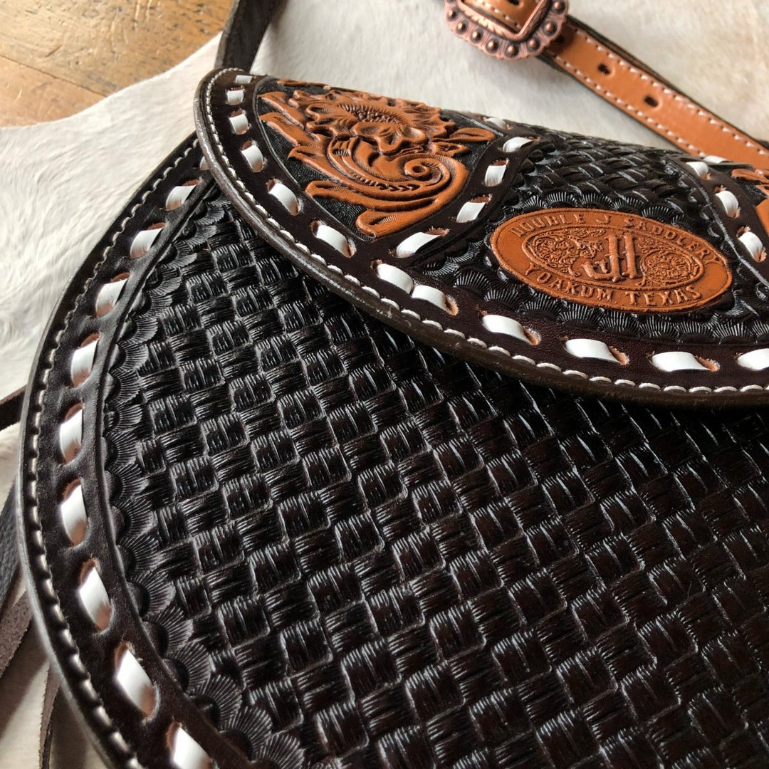 Close-up of The Mattawa saddle bag by Savannah Sevens western life{&}style, featuring intricate basket weave and floral patterns with a stamped oval label. The edge is accented with white leather lacing. By Double J. 
