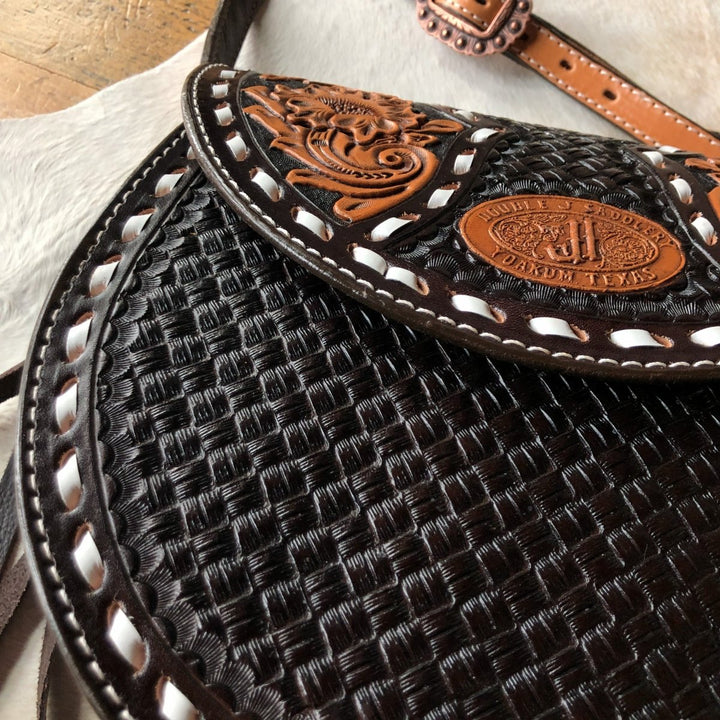 Close-up of The Mattawa saddle bag by Savannah Sevens western life{&}style, featuring intricate basket weave and floral patterns with a stamped oval label. The edge is accented with white leather lacing. By Double J. 