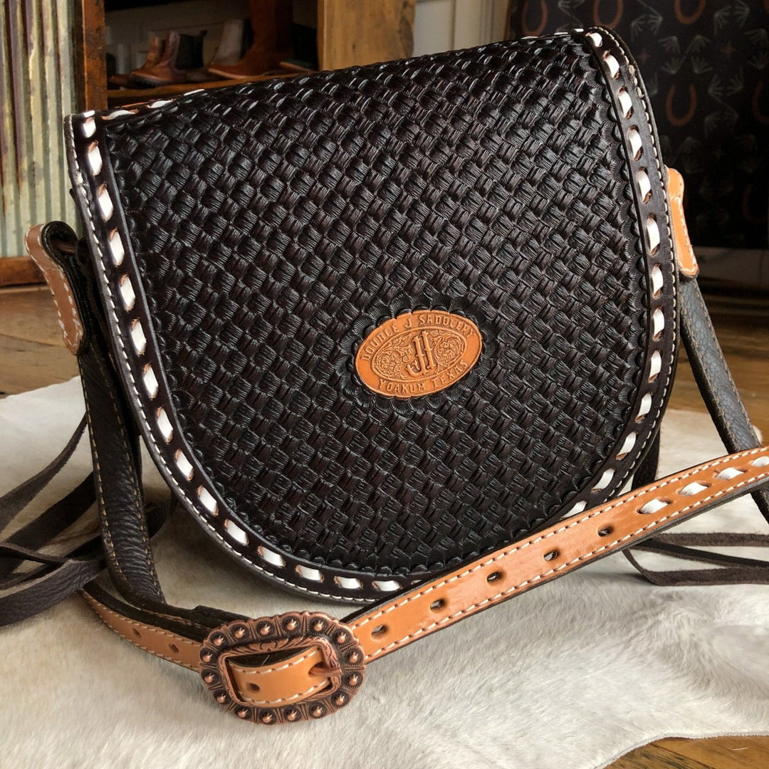 The Mattawa by Savannah Sevens western life{&}style is a handcrafted dark brown leather saddle bag with a basket weave pattern, tan trim, and a western-style buckle, displayed on a light cowhide rug.