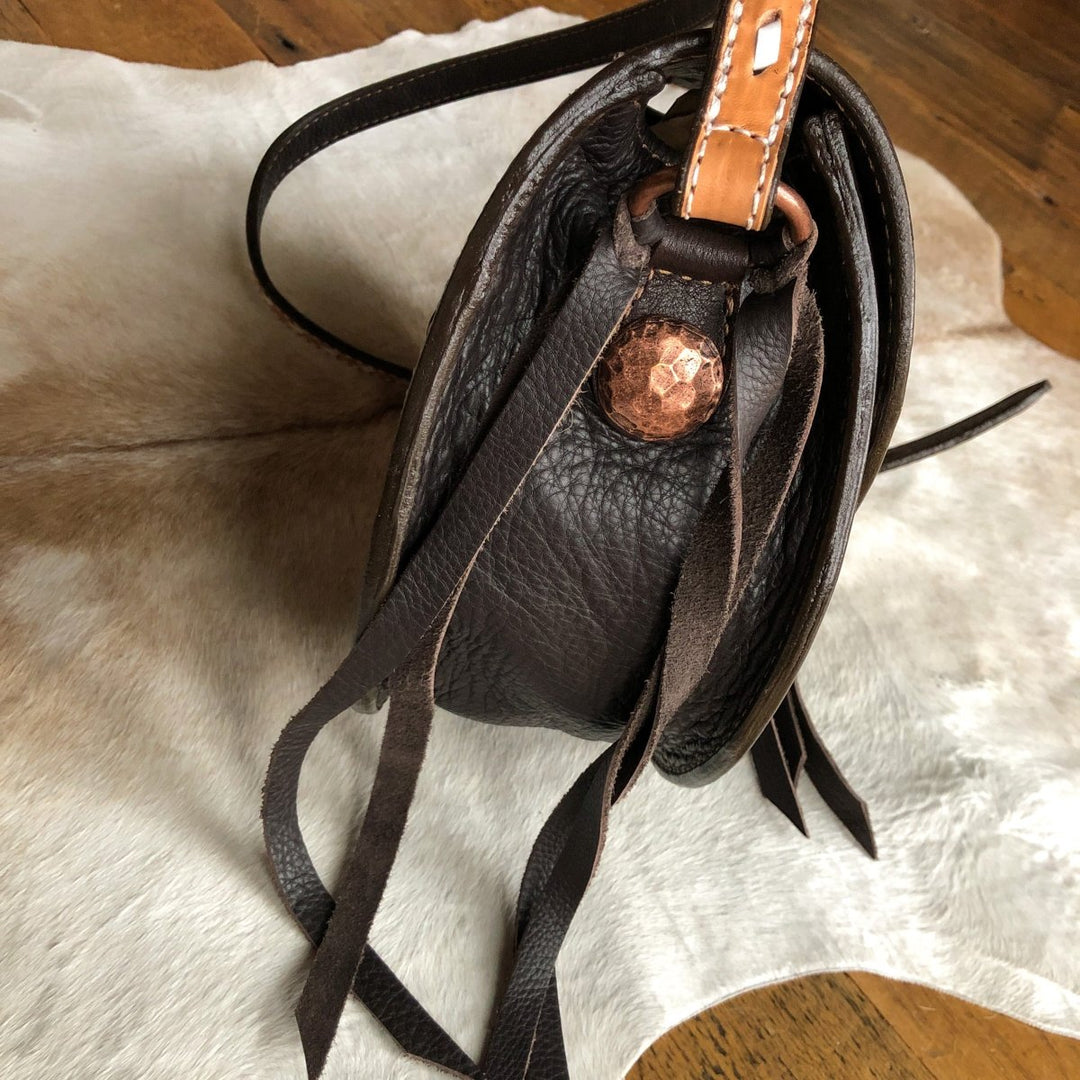 The Mattawa saddle bag by Savannah Sevens western life{&}style features dark brown leather with basket weave texture, a copper button, and long fringe, shown on an animal hide rug over a wooden floor.