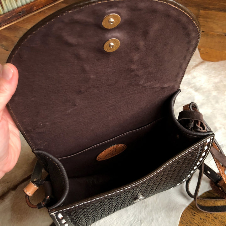 A hand opens the flap of The Mattawa by Savannah Sevens western life{&}style, a black basket weave leather saddle bag with brown lining and snap closures, showing its empty interior and a small stitched Double J Saddlery logo patch.