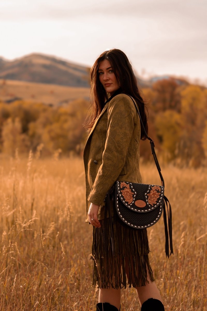 A woman stands in a sunlit field wearing a green jacket and fringe skirt, holding The Mattawa saddle bag from Savannah Sevens western life{&}style. Autumn trees and hills form a picturesque background. By Double J. 