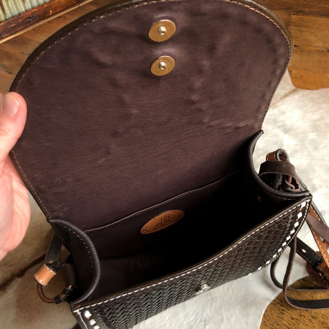 A hand opens the flap of The Mattawa by Savannah Sevens western life{&}style, a black basket weave leather saddle bag with brown lining and snap closures, showing its empty interior and a small stitched Double J Saddlery logo patch.