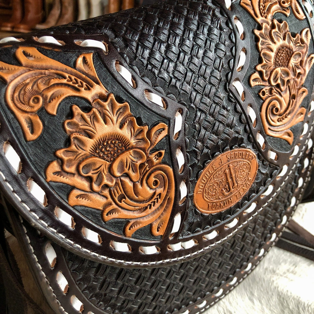 Close-up of The Mattawa saddle bag by Savannah Sevens western life{&}style, showcasing intricate floral and basket weave patterns, a brown oval label with engraved text, and white stitching along the edges. By Double J. 