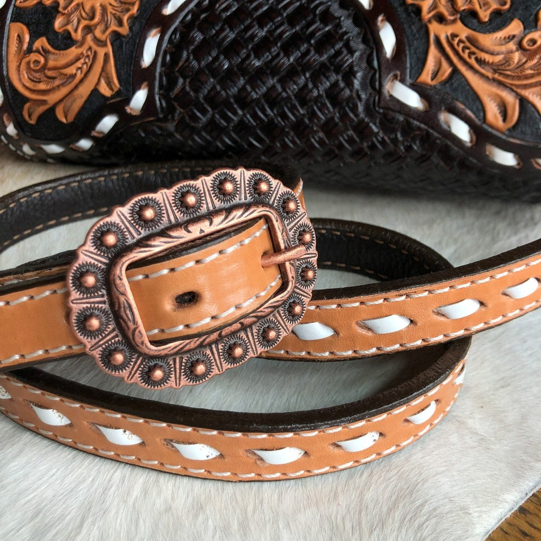 A close-up of The Mattawa, a brown leather belt with white accents and an ornate bronze buckle by Savannah Sevens western life{&}style, placed on light fur with a basket weave saddle bag in the background. By Double J. 