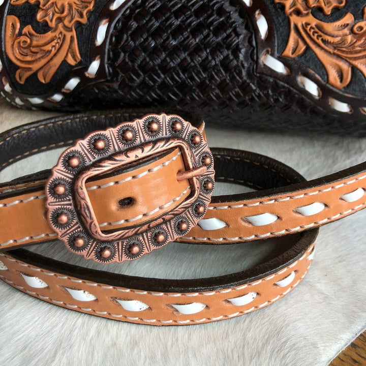 A close-up of The Mattawa, a brown leather belt with white accents and an ornate bronze buckle by Savannah Sevens western life{&}style, placed on light fur with a basket weave saddle bag in the background. By Double J. 