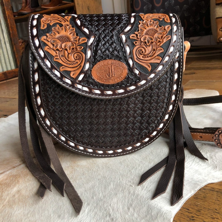 The Mattawa by Savannah Sevens western life{&}style is a handcrafted leather saddle bag featuring floral carvings, basket weave patterns, white stitching, a round silver emblem, and fringe accents, displayed on a hide rug. By Double J. 