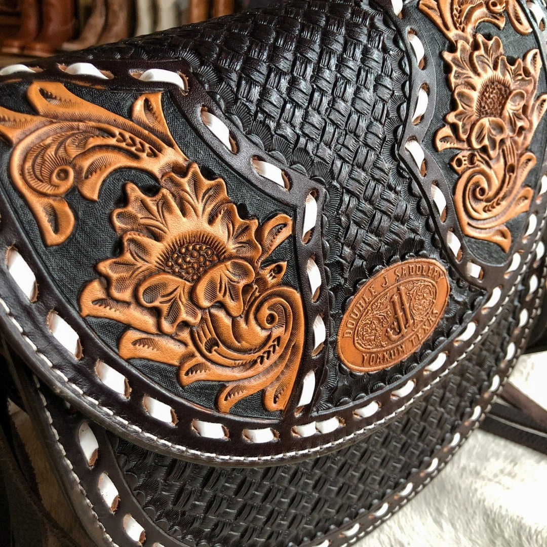 Close-up of The Mattawa saddle bag by Savannah Sevens western life{&}style, showcasing intricate floral and basket weave patterns, a brown oval label with engraved text, and white stitching along the edges. By Double J. 