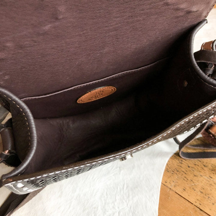 The Mattawa by Savannah Sevens western life{&}style is an open brown leather saddle bag with basket weave detailing, a stitched leather tag on the inside pocket, shown on a wooden surface against a white textured background. By Double J. 