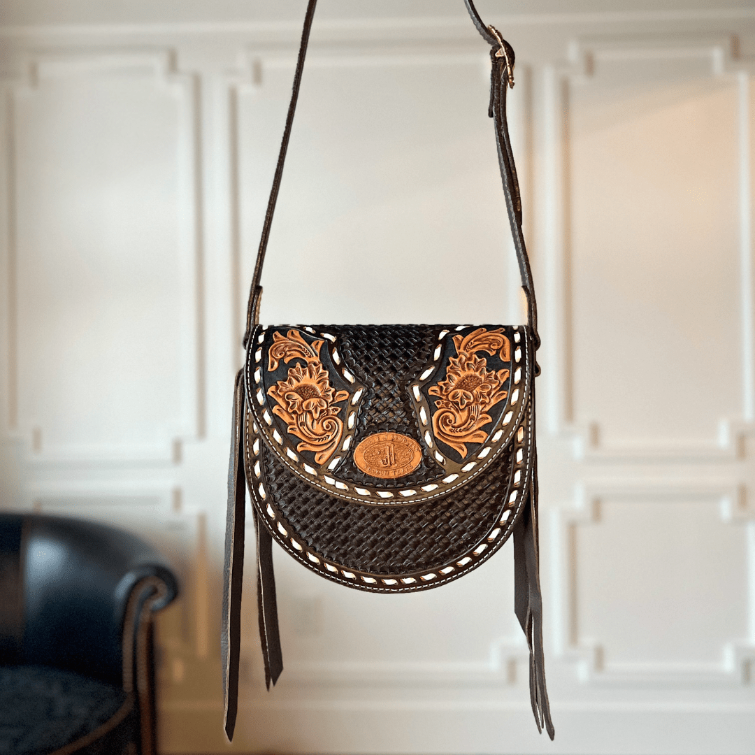 The Mattawa by Savannah Sevens western life{&}style is a black leather saddle bag featuring tan floral tooling, basket weave details, and white stitching, displayed against a white paneled wall with a dark chair partly visible in the background. By Double J. 