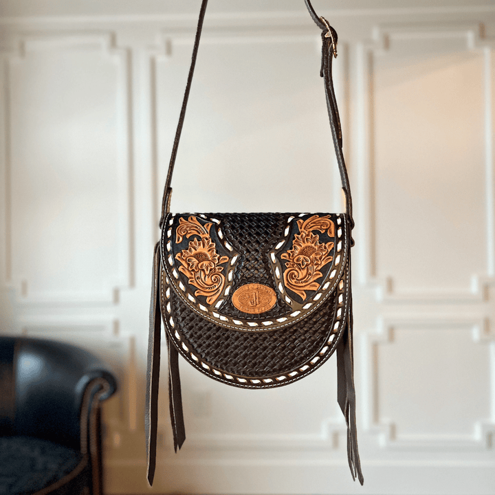 The Mattawa by Savannah Sevens western life{&}style is a black leather saddle bag featuring tan floral tooling, basket weave details, and white stitching, displayed against a white paneled wall with a dark chair partly visible in the background. By Double J. 