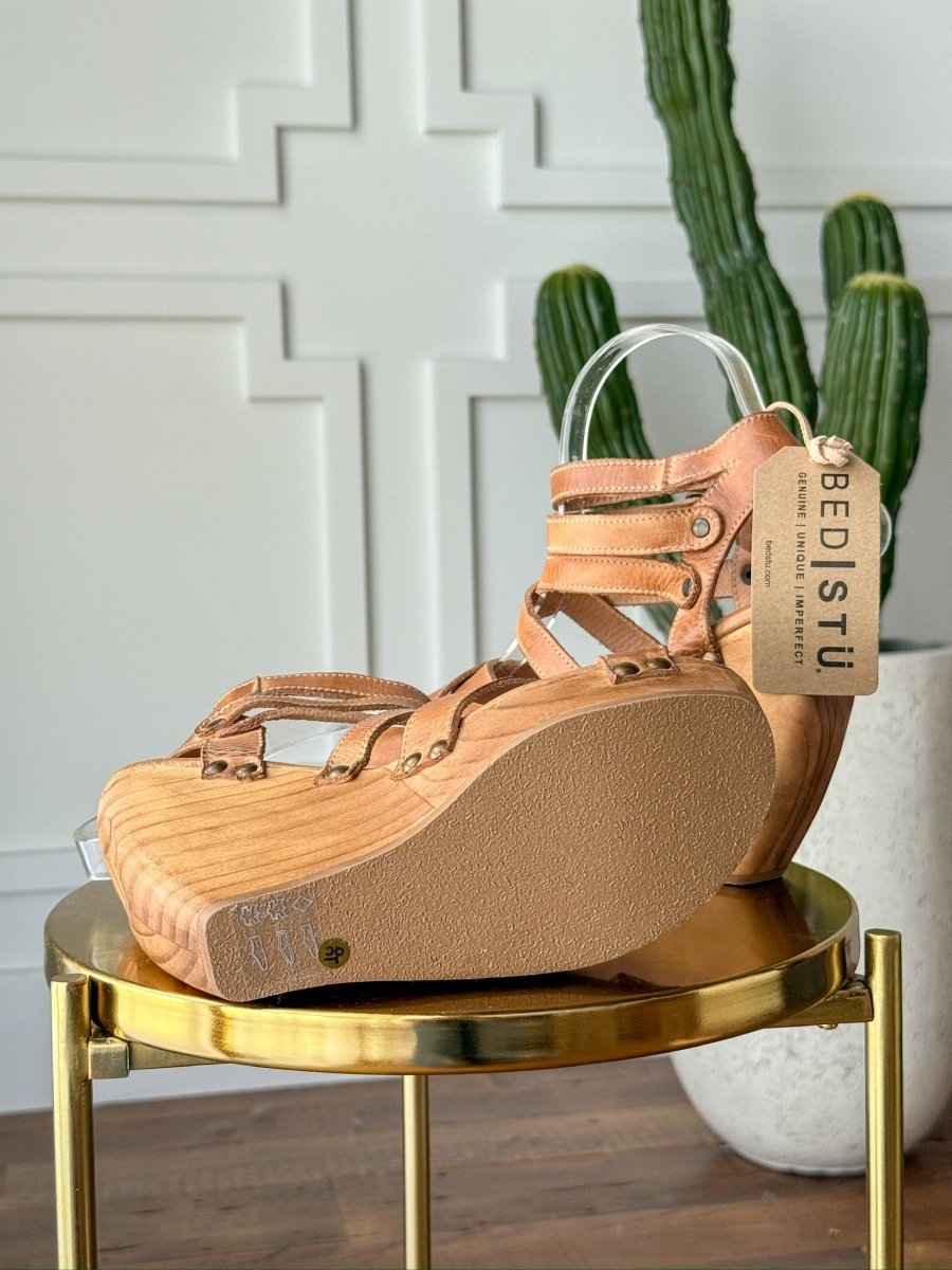 The Mesilla by Savannah Sevens western life{&}style, a tan leather strappy heel with open toe, sits on a round gold table. A tall green cactus and white paneled wall are in the background. Made by BedStu. 