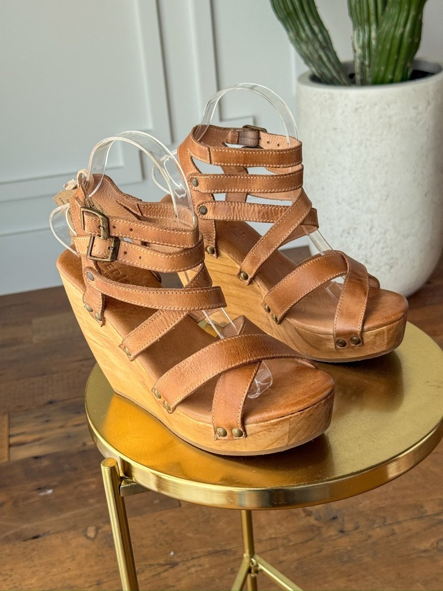 The Mesilla by Savannah Sevens western life{&}style, tan leather wedge sandals with multiple straps and gold buckle detail, rest on a round gold table with a wooden floor, white planter, and green cactus in the background. Made by BedStu. 