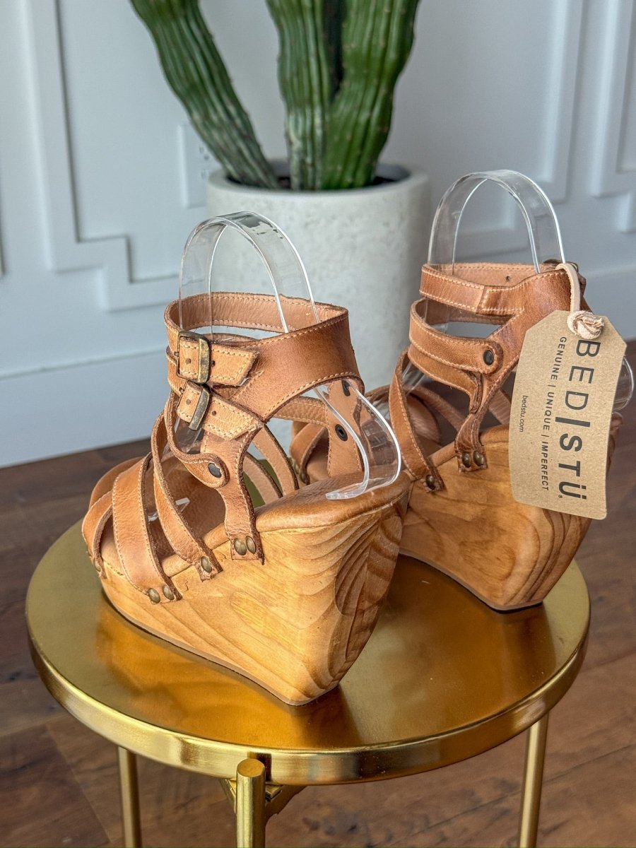A pair of The Mesilla tan leather wedge sandals by Savannah Sevens western life{&}style, featuring wooden platform heels and buckle straps, are displayed on a round golden table with a tall potted cactus in the background. Made by BedStu.