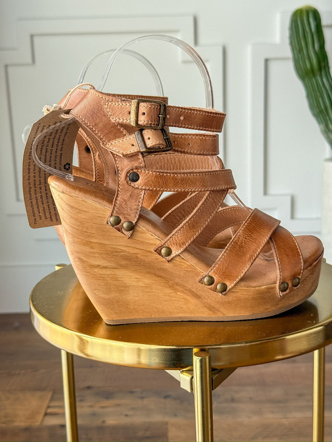The Mesilla by Savannah Sevens western life{&}style is a tan leather sandal with multiple straps, metal buckles, and brass studs, featuring an open toe and high wooden platform heel. It's displayed on a round gold table with a cactus in the background. Made by BedStu. 