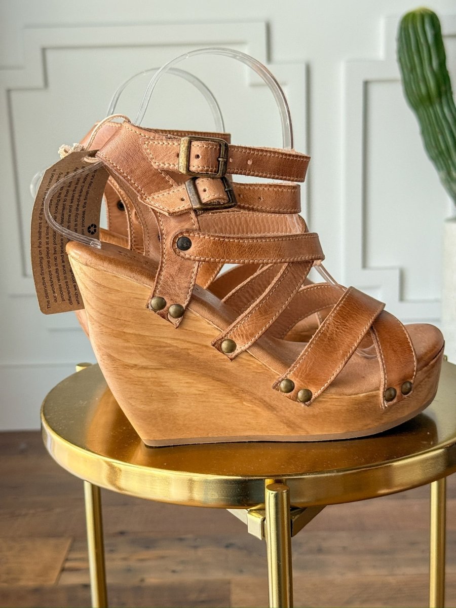 The Mesilla by Savannah Sevens western life{&}style is a tan leather sandal with multiple straps, metal buckles, and brass studs, featuring an open toe and high wooden platform heel. It's displayed on a round gold table with a cactus in the background. Made by BedStu. 
