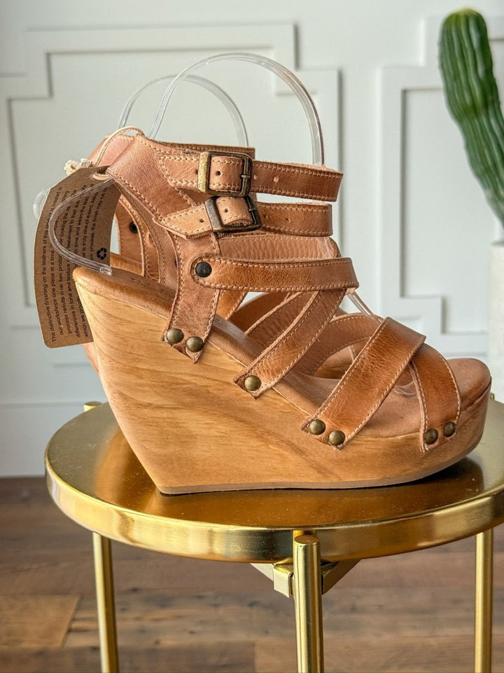 The Mesilla by Savannah Sevens western life{&}style is a tan leather sandal with multiple straps, metal buckles, and brass studs, featuring an open toe and high wooden platform heel. It's displayed on a round gold table with a cactus in the background. Made by BedStu. 