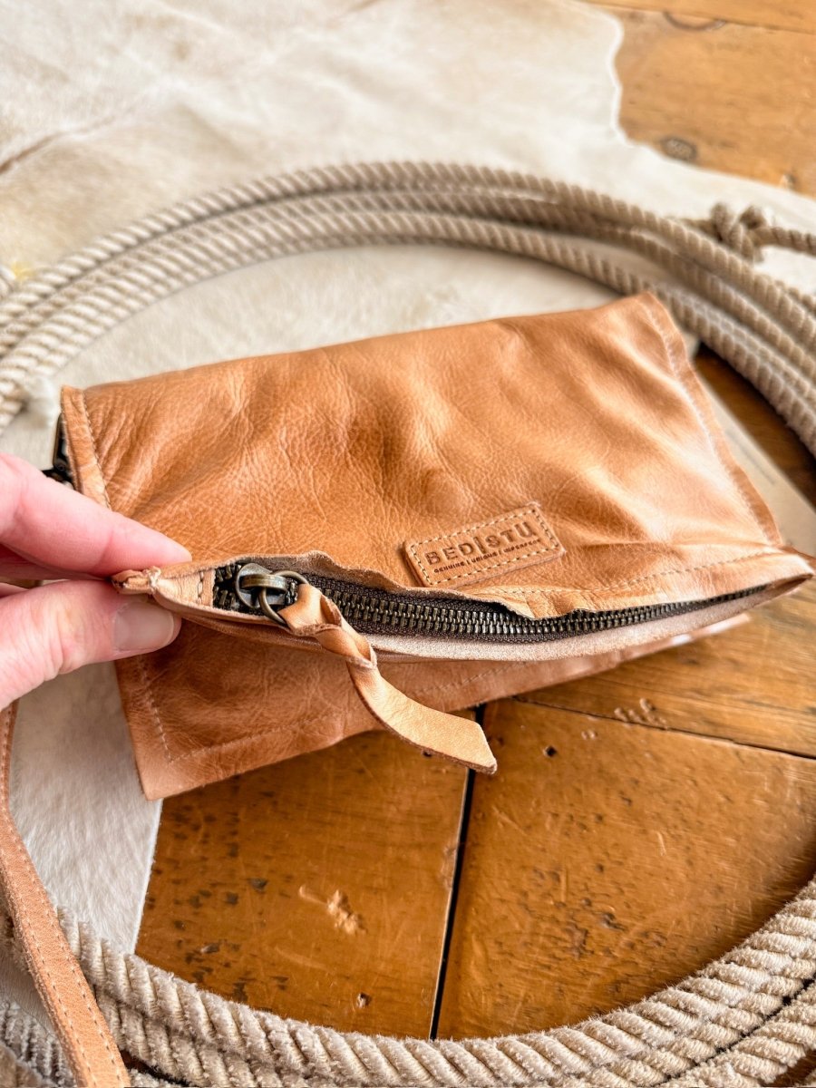 A hand opens the zipper of The Mineola leather clutch by Savannah Sevens western life{&}style. The small, light brown pouch rests on wood, with a coiled rope and animal hide beneath it.