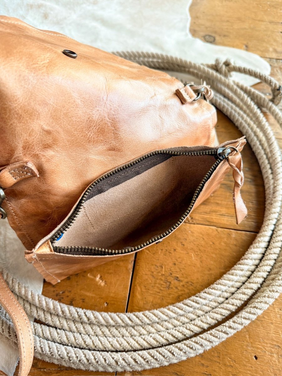 The Mineola by Savannah Sevens western life{&}style, a tan leather crossbody with a zippered opening, rests on a coiled rope atop wood and a light hide. The bag is partially open, showing its suede interior.