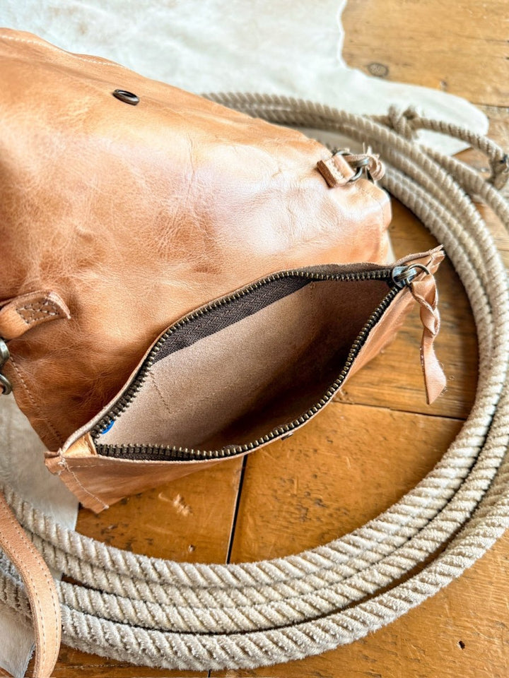 The Mineola by Savannah Sevens western life{&}style, a tan leather crossbody with a zippered opening, rests on a coiled rope atop wood and a light hide. The bag is partially open, showing its suede interior.