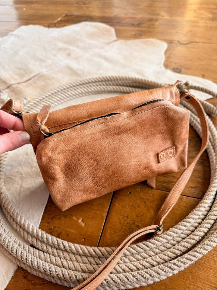 The Mineola by Savannah Sevens western life{&}style—a tan leather crossbody bag with a zipper—is held by a hand, resting on coiled rope and light fabric atop a wooden floor.