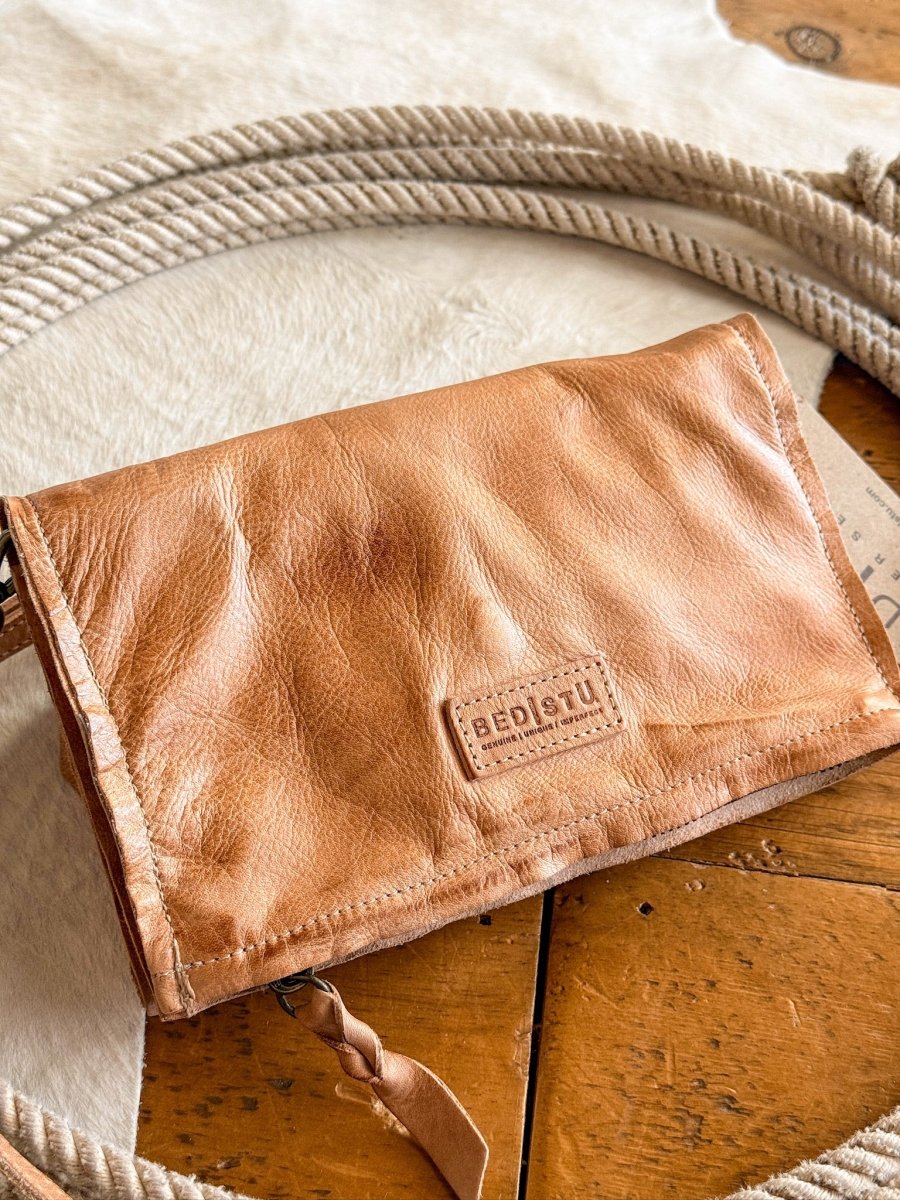 The Mineola crossbody by Savannah Sevens western life{&}style rests on a wooden surface, with a coiled rope and light cowhide rug nearby.