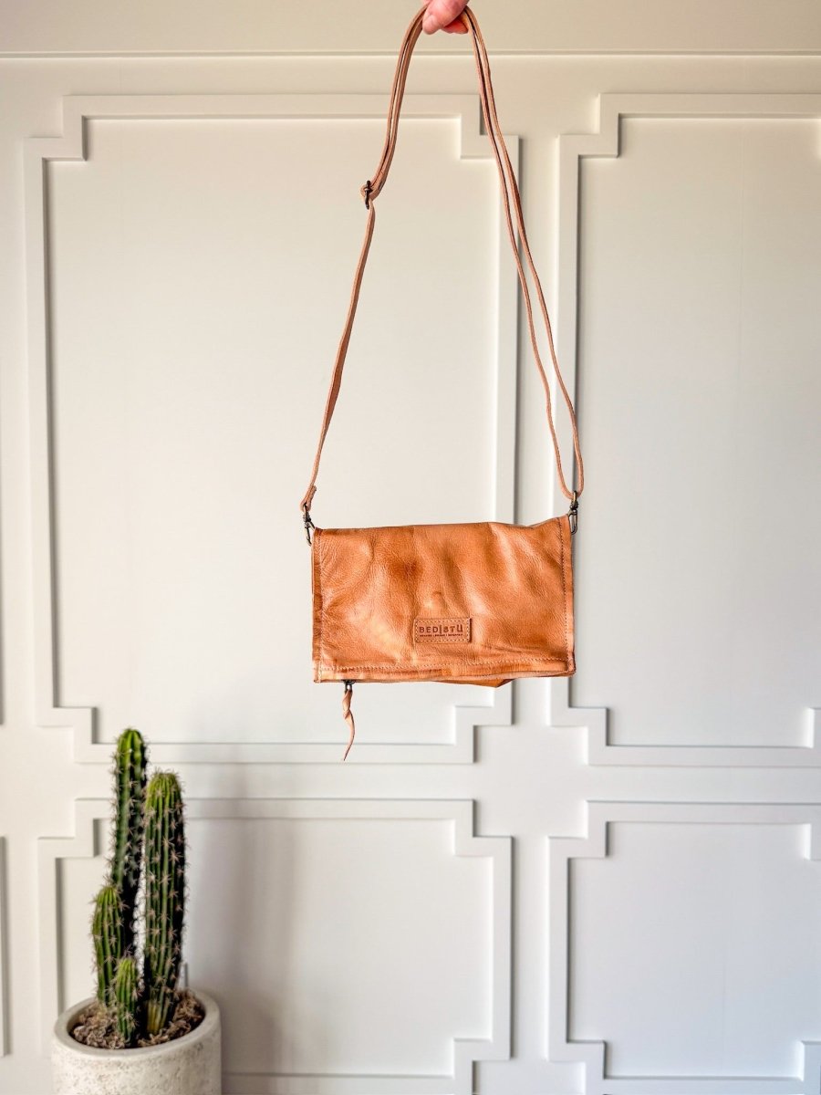 A hand holds up The Mineola tan leather crossbody bag by Savannah Sevens western life{&}style, featuring a long strap, in front of a beige paneled wall; a potted cactus sits on the floor in the bottom left corner.