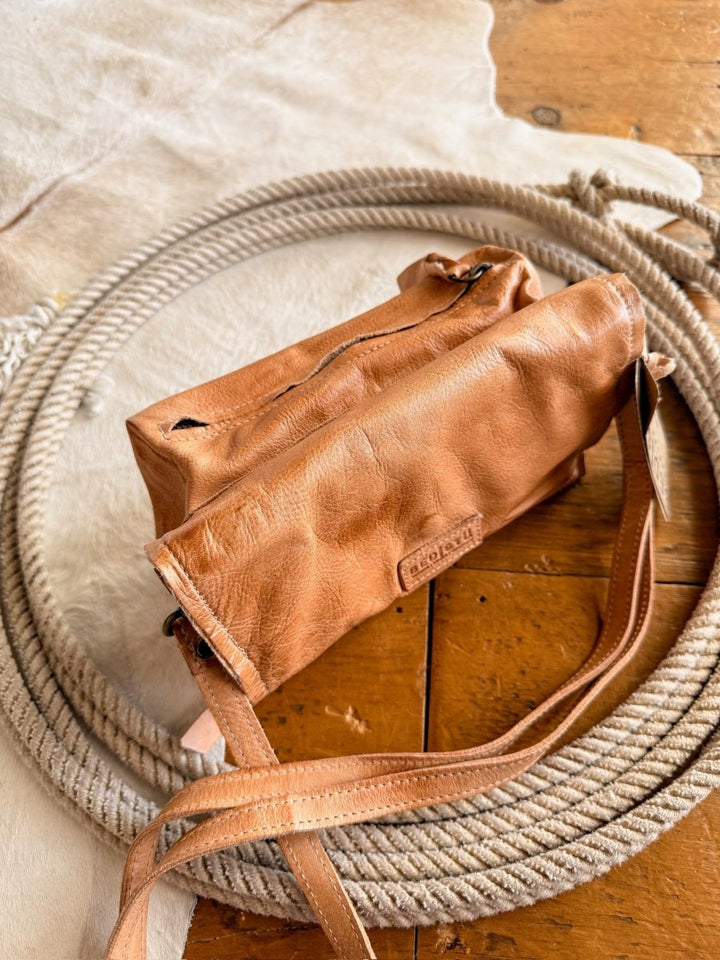 The Mineola by Savannah Sevens western life{&}style is a tan leather crossbody bag featuring a zipper, displayed on coiled rope atop cowhide and wood, exemplifying the brand’s signature craftsmanship.