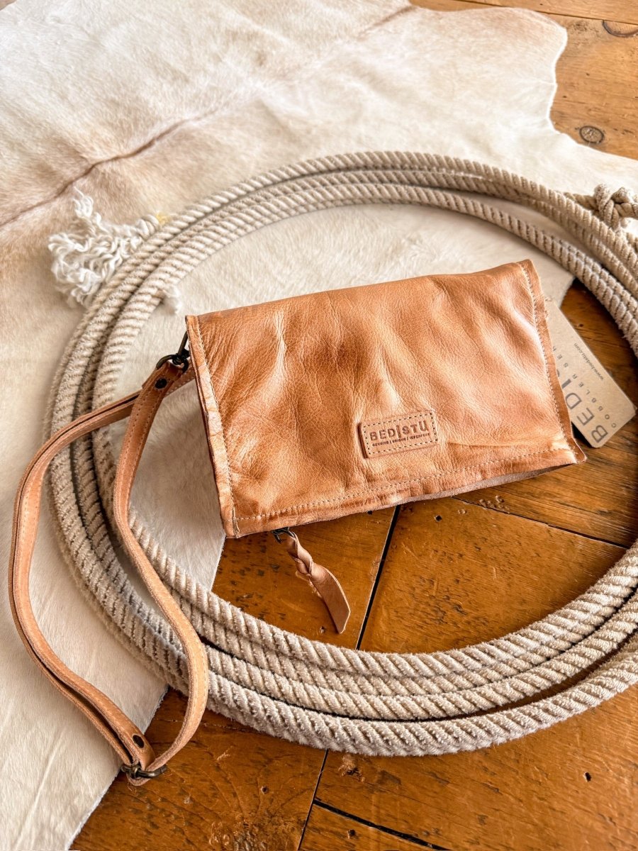 The Mineola wristlet by Savannah Sevens western life{&}style, crafted in tan leather with a logo, rests on a white rug over wood. A small product tag is partly visible next to the dip-dye leather clutch.