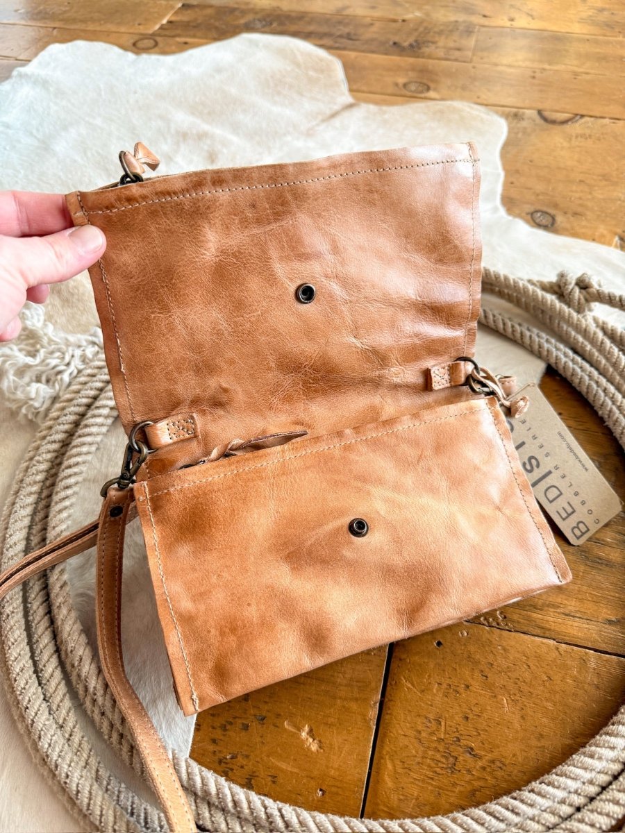A hand holds open The Mineola, a tan leather crossbody bag by Savannah Sevens western life{&}style, showing its inner compartment. The bag rests on a wooden floor near a coiled rope and its signature tag.