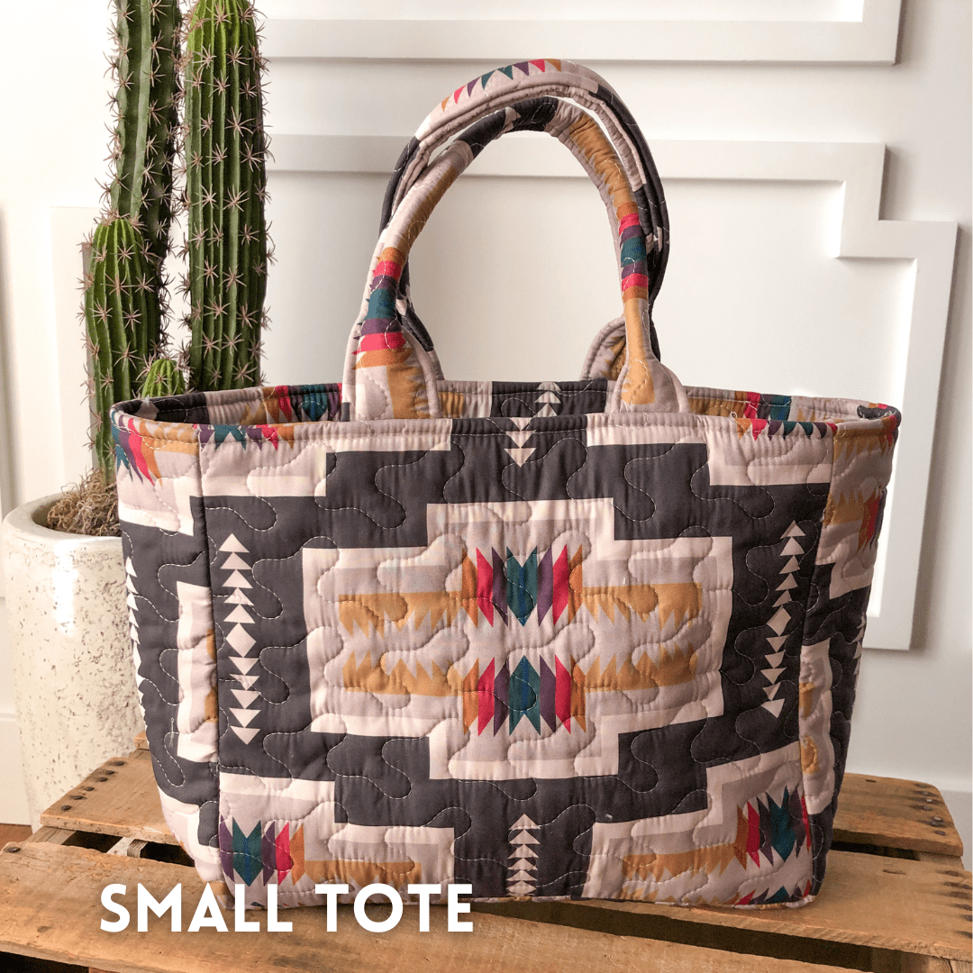 A quilted bag from The Muskogee Collection by Savannah Sevens western life{&}style, featuring colorful geometric patterns in beige, black, and red, rests on a wooden surface beside a potted cactus; SMALL TOTE appears in the lower left. By Ida Mae. 